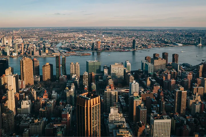 大都市の広大な空撮。手前に多数の高層ビルが密集し、その奥に広い川が街を横断している。川の両岸にはさらに多くの建物が広がり、遠くまで都市の景観が続いている。夕暮れ時の柔らかな光が街を照らしている。