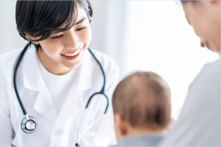 白衣を着た女性医療従事者が笑顔で赤ちゃんを見つめている。手前には赤ちゃんの頭部がぼやけて写っており、右側には別の人物の肩と頭部が一部見える。明るく清潔感のある医療現場の様子。