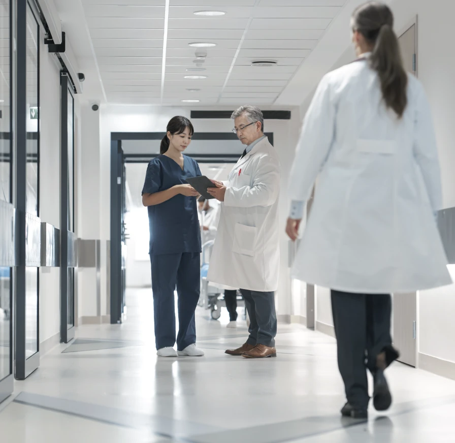 診察室で男性医師が女性患者に説明している様子。医師はダークブルーのスクラブを着用し、笑顔で右手を広げて話しており、患者は穏やかな表情で耳を傾けている。二人の間には医療情報が表示された大きなモニターが置かれ、背景には大きな窓から自然光が差し込んでいる。