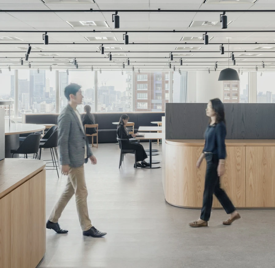 明るい会議室で5人のビジネスパーソンが笑顔で打ち合わせをしている様子。左端に立つ男性はダークグレーのジャケットと白いTシャツを着用し、腕を組んで右方向を見て微笑んでいる。中央に立つ男性は白い長袖シャツを着用し、両手を前で組んで、笑顔で参加者全体を見渡している。テーブルには3台のノートパソコンが置かれ、座っている3人の男女は皆笑顔で、中央の男性の方を見ている。左から2番目の女性は水色のシャツを着て、ノートパソコンの前に座っている。中央の男性の右隣に座る女性はベージュのジャケットを着用し、右端に座る男性はネイビーのスーツを着ている。全体的に活気があり、ポジティブな雰囲気の会議風景。